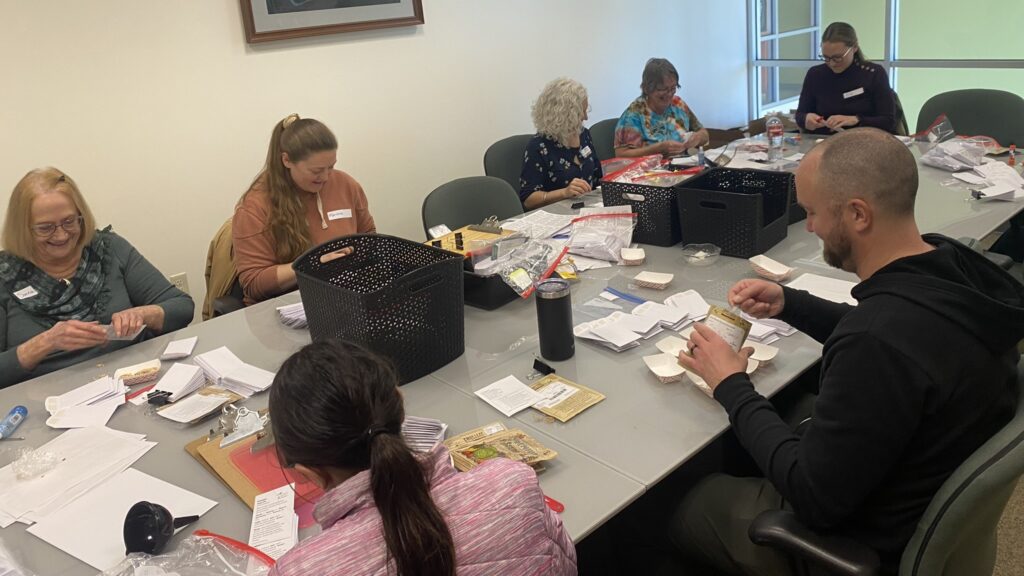 Seven Seed Library Volunteers sitting at a conference table packaging seeds
